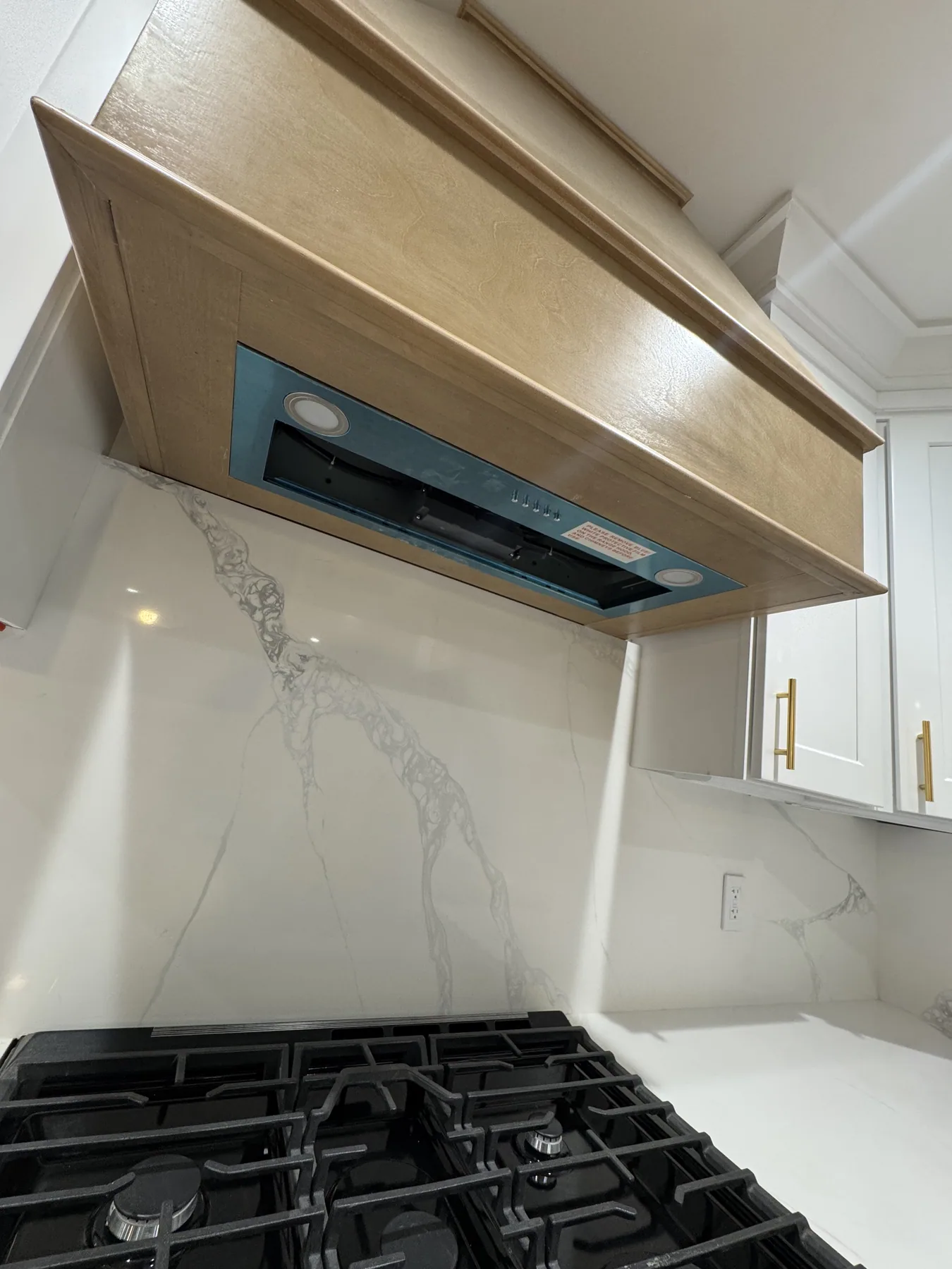 Custom wood range hood above a gas cooktop with a marble backsplash and white cabinets
